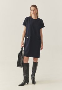 Navyblå shiftklänning i slät tyg, med korta ärmar och två framfickor med detaljer i guldknapp. Modellen håller en svart tote-väska.