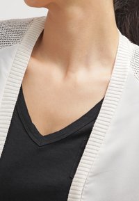 Kvinna som bär en svart V-ringad tröja och en ljusbeige cardigan med ribbad kant, vilket visar hennes hals och övre bröst.