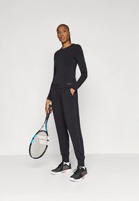 Γυναίκα που στέκεται φορώντας μαύρο μπλουζάκι μακρύ μανίκι και παντελόνι, κρατώντας μία ρακέτα τένις, με μαύρα και άσπρα αθλητικά παπούτσια, σε λευκό φόντο.