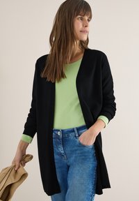 Femme aux longs cheveux bruns portant un cardigan noir sur un haut vert clair et un jean bleu, tenant une veste beige dans une main.