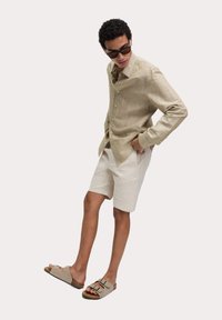 Lichtbruin gestreept shirt met lange mouwen, beige linnen shorts en bruine slip-on sandalen. De outfit is lichtgewicht en casual.