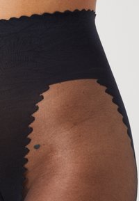 Schwarze hoch taillierte Strumpfhosen mit transparenten Details, die mit gewellten Kanten an der Taille und den Beinöffnungen versehen sind und die Haut darunter betonen.