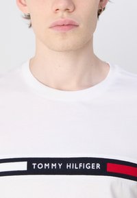 Valge puuvillane T-särk, millel on horisontaalne tumesinine ja punane triibuline riba ning rinnal valgete tikanditega tekst "TOMMY HILFIGER".