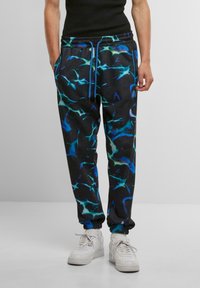 Sorte joggerbukser med et blåt og teal abstrakt mønster, elastisk talje med snor og afsluttede ben. Båret med hvide sneakers.