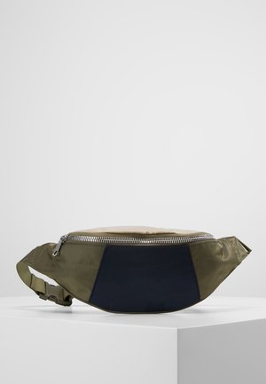 Petit sac banane bicolore noir et vert olive en tissu avec fermeture éclair argentée et sangle réglable sur surface blanche.