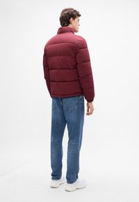 Bordeaux pufferjakke med vandret syninger, høj krave og elastiske manchetter, parret med lyseblå jeans og hvide sneakers.