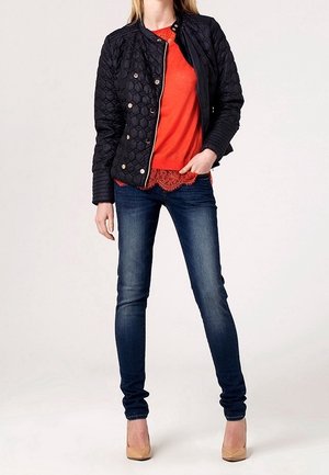 Frau trägt eine schwarze gesteppte Jacke, rote Bluse mit Spitzenbesatz, dunkle enge Jeans und beige High Heels, steht mit der Hand in der Tasche.