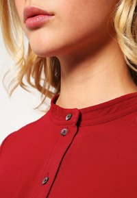 Blouse rouge avec un col rond, ornée d'une bordure texturée et de deux boutons sombres au niveau du col. Tissu lisse, vue rapprochée du vêtement supérieur.
