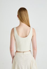 Donna con capelli lunghi e lisci che indossa un top corto senza maniche beige e una gonna alta beige chiara, in piedi contro uno sfondo bianco.