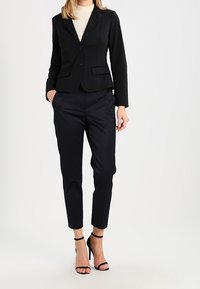 Czarna marynarka z dwiema przednimi kieszeniami, zestawiona z teksturowanymi ciemnymi spodniami. Modelka ma na sobie czarne sandały na obcasie oraz jasny top pod marynarką.