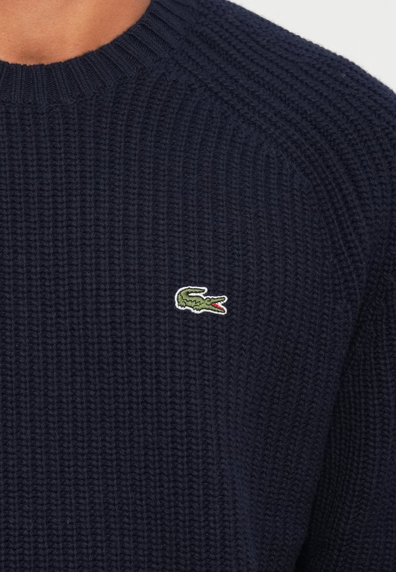 Marinblått stickat tröja med ribbat mönster, som har en liten grön krokodil-logotyp på bröstet. Rund halsringning, avslappnad passform.