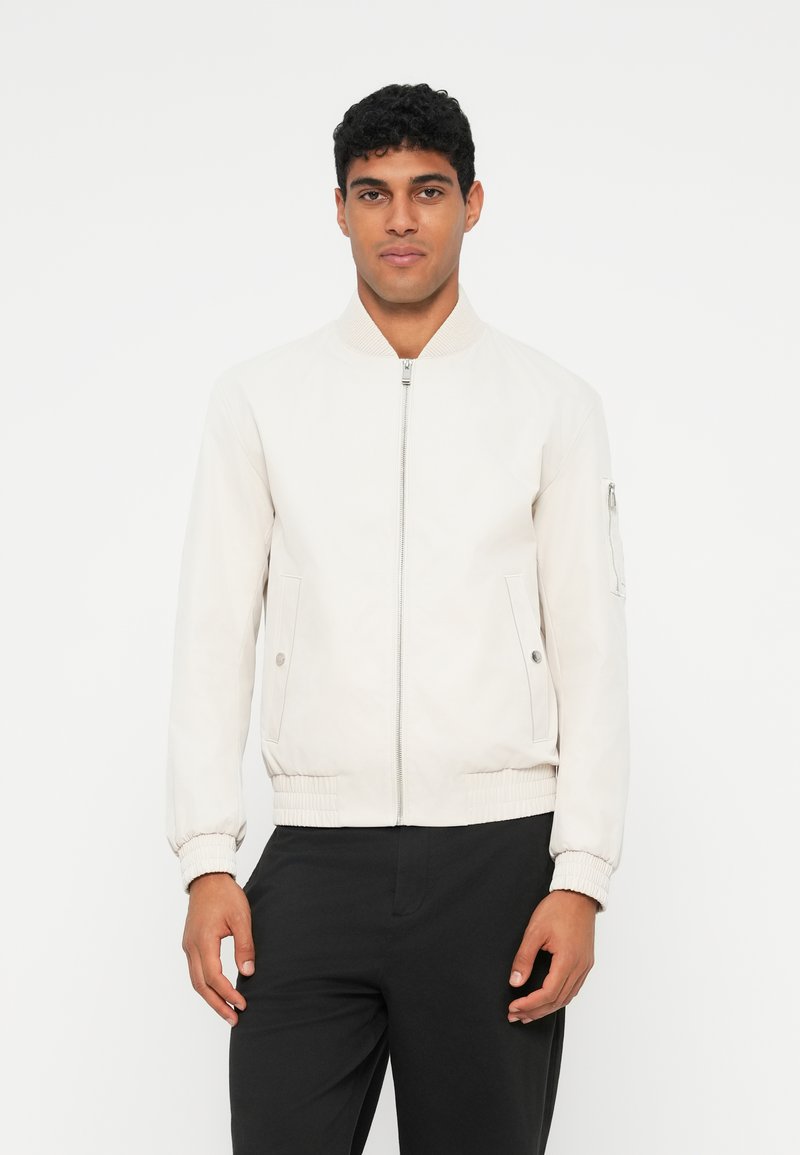 Corta-vento bege claro com fecho de correr à frente, gola e punhos canelados, e bolsos laterais. Textura suave, design minimalista e sem padrões visíveis.