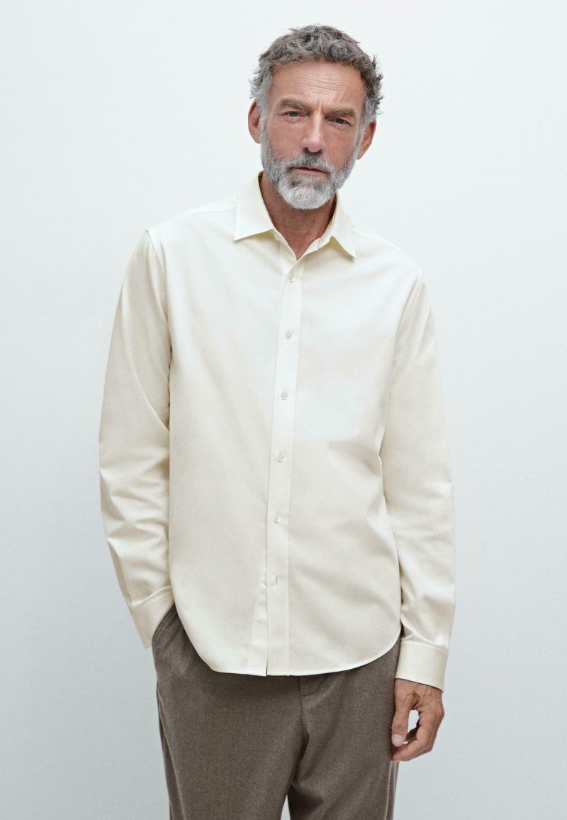 Camicia button-up color crema con colletto classico, maniche lunghe e una texture liscia, abbinata a pantaloni marrone chiaro.