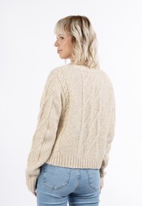 Beige kötött pulóver kábel és textúrált mintákkal. Bordás szegéllyel, hosszú ujjakkal és laza fazonnal rendelkezik, kék farmerrel párosítva.
