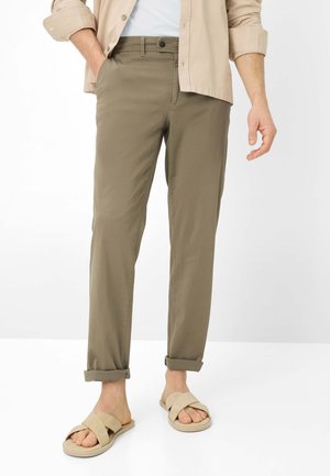 Mann trägt hochgekrempelte olivgrüne Hose, beige Sandalen mit offenen Zehen, weißes Hemd und eine beige Langarmjacke, die Hände entspannt.