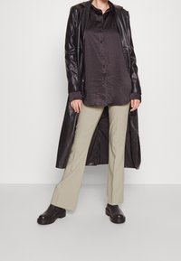 Zwarte leren lange jas over een donker button-up shirt, gecombineerd met lichtbeige uitlopende broek en zwarte enkellaarsjes. Gladde texturen, contrasterende kleuren.