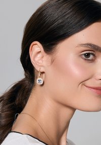 Złote kolczyki w kształcie obręczy z okrągłym wzorem, w centrum z niebieskim kamieniem otoczonym mniejszymi przezroczystymi kamieniami.