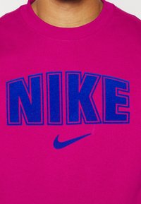 Felpa magenta con lettering blu in grassetto "NIKE" e un piccolo logo swoosh Nike blu. Materiale in cotone con una texture morbida.