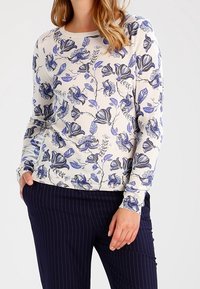 Frau, die einen weißen Pullover mit blauen Blumenmustern und dunkelblauen gestreiften Hosen trägt, steht mit einer Hand in der Tasche.