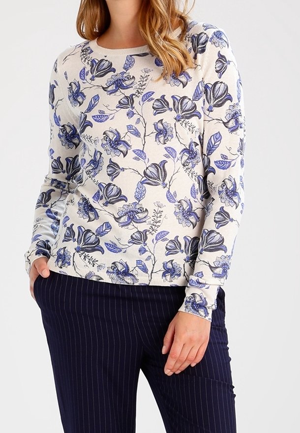 Frau, die einen weißen Pullover mit blauen Blumenmustern und dunkelblauen gestreiften Hosen trägt, steht mit einer Hand in der Tasche.