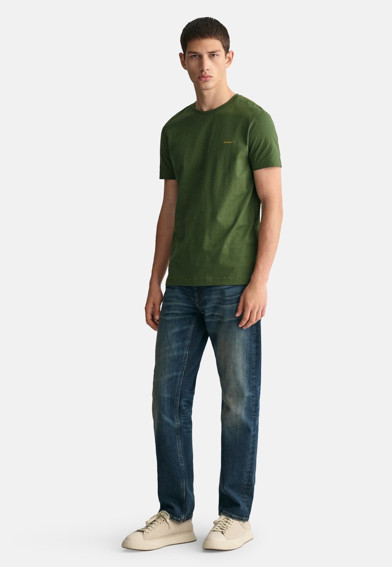 Jongeman staat rechtop, draagt een groen t-shirt met korte mouwen, blauwe jeans en beige sneakers, kijkt recht vooruit met een neutrale uitdrukking.