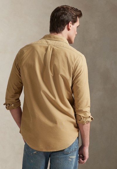 Ανοιχτό καφέ πουκάμισο με μακριά μανίκια, κατασκευασμένο από απαλό ύφασμα, με κυρτή γραμμή στο τελείωμα, κουμπωτά μανσέτα και πλάγιες πιέτες στην πλάτη. Φορεμένο με καταστραμμένο μπλε τζιν.