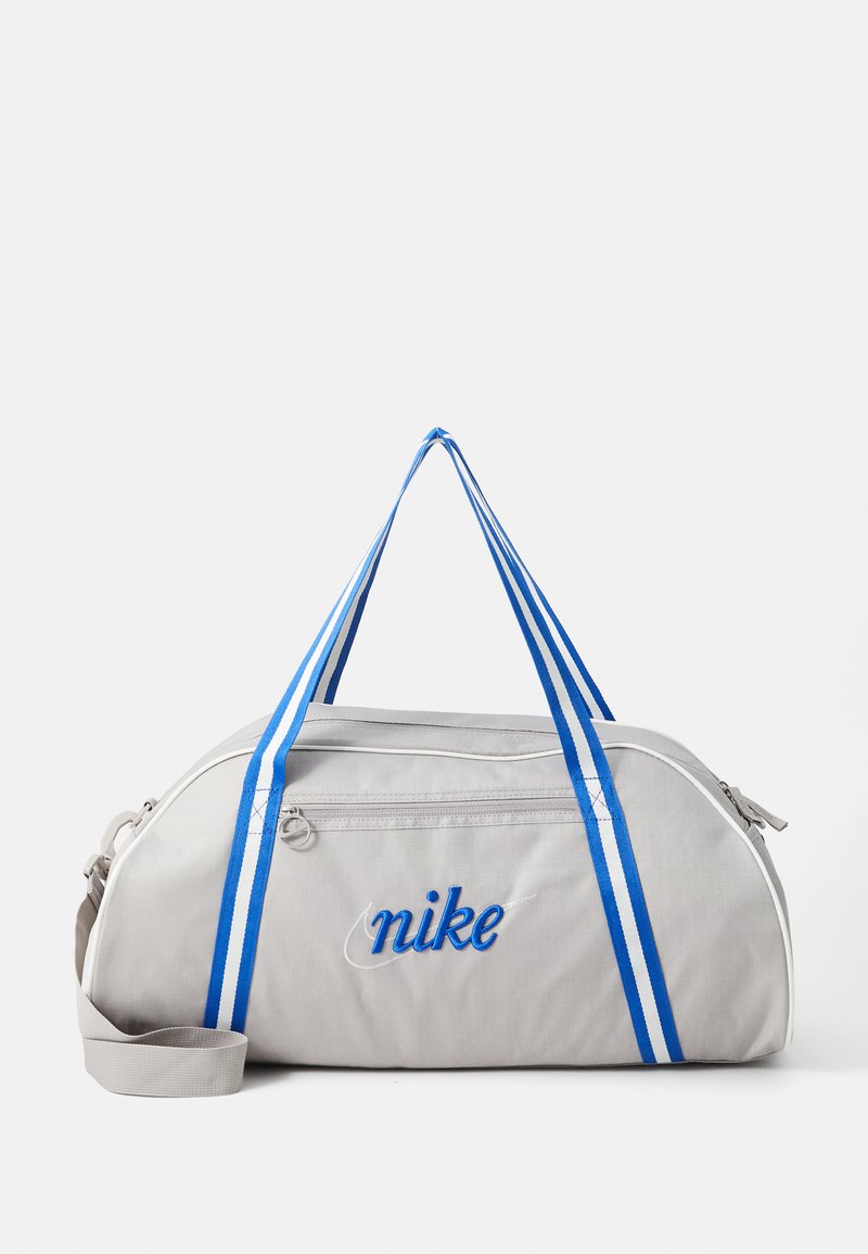 Siva torba za duffel s plavim trakama, sa zatvaranjem na zatvarač i izvezenim logotipom "Nike". Izrađena od izdržljivog materijala s glatkom teksturom.