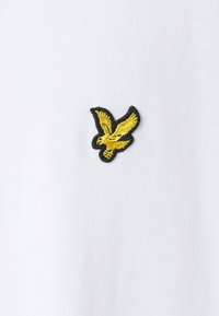 Hvidt stof med et sortkantet gult broderet fugleemblem. Fuglen har detaljerede vinger og en strømlinet form.