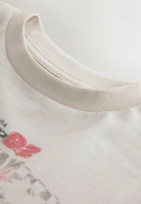 Prim-plan al unui tricou bej cu guler rotund, evidențiind cusătura fină a gulerului tip dantelă și un design parțial imprimat, estompat, cu text în roșu și negru.