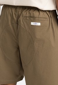 Braune Shorts aus leichtem Stoff mit strukturierter Oberfläche. Mit elastischem Bund, einer Gesäßtasche und einem runden Stoffdetail.