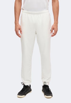 Uomo che indossa pantaloni da jogging bianchi con caviglie elastiche e sneakers nere su sfondo neutro.