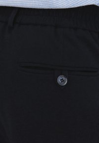 Czarne spodnie z elastycznym pasem, z przednią kieszenią z płaskim zapięciem i szarym guzikiem. Gładka faktura materiału.