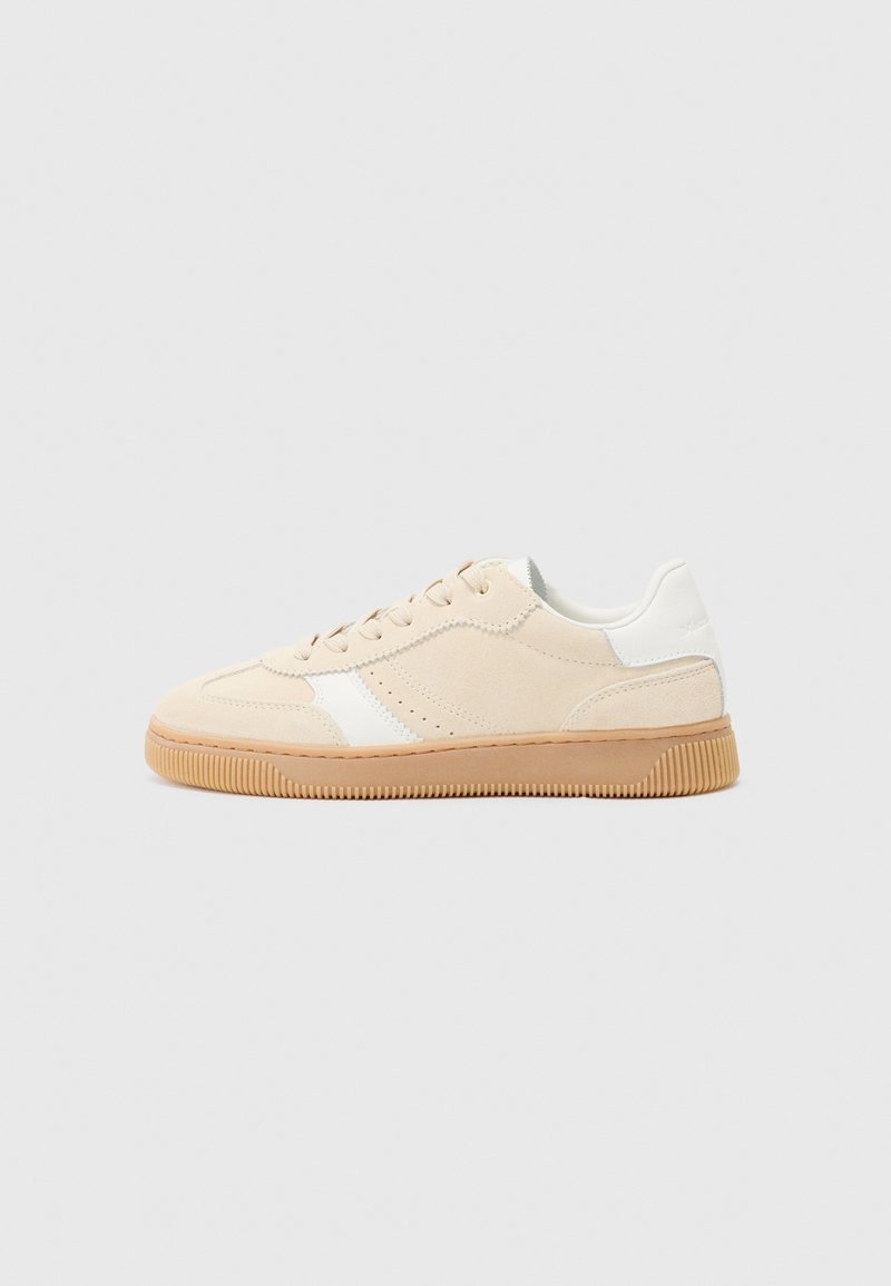 Baskets basse beige avec accents blancs et semelle en gomme, présentées de côté sur un fond gris clair.