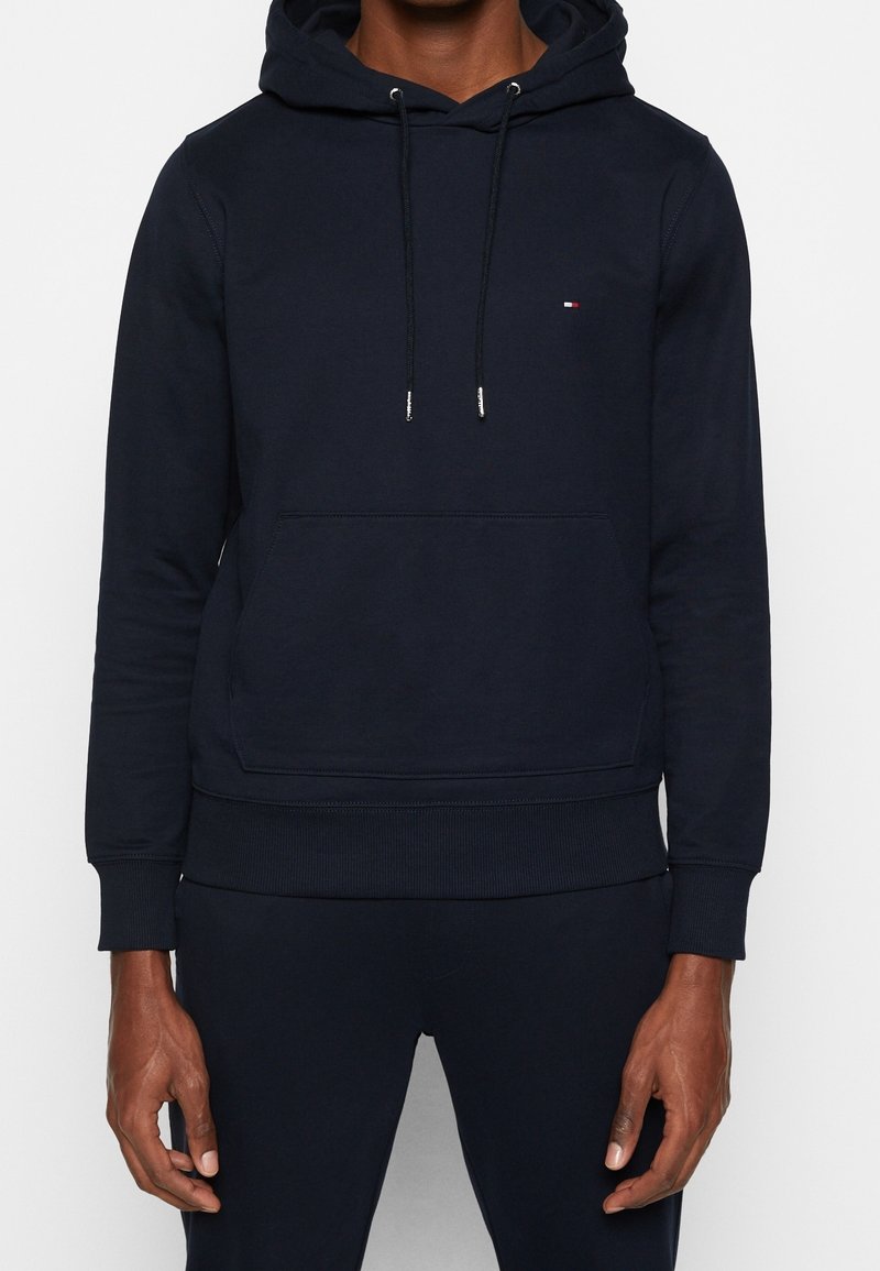 Homme portant un sweat à capuche bleu marine avec poche avant et petit logo rouge, blanc et bleu sur la poitrine, debout, bras détendus le long du corps.