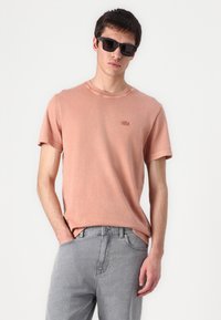 Kortermet, ferskenfarget bomull T-skjorte med rund hals og brodert logo. Kombinert med lysegrå denimjeans.