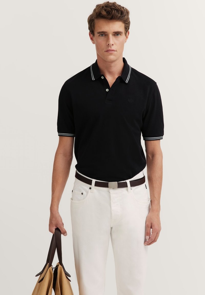 Schwarzes Polo-Shirt mit weißen Kragenstreifen, kurze Ärmel. Kombiniert mit weißen Jeans und einem braunen Gürtel. Hält eine sandfarbene Tasche. Einfaches Design.