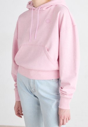 Personne portant un sweat à capuche rose clair avec poche avant et cordons, associée à un jean bleu clair sur un fond uni.