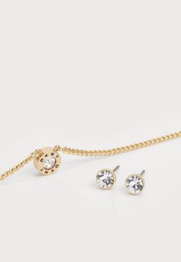 Gold chain bracelet featuring a central circular charm with a clear crystal. Paired with two gold-stud earrings, each set with a clear stone.