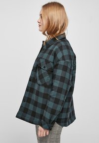 Camicia di flanella a quadri verde e nera con chiusura a bottoni, dotata di due tasche sul petto e dalla vestibilità comoda, indossata sopra pantaloni a scacchi.
