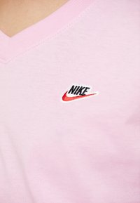 Ljust rosa bomullströja med V-ringning och en svart och röd Nike-logotyp broderad på bröstet. Slät textur.