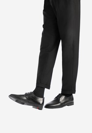 Des chaussures en cuir noir au fini lisse, dotées de lacets et de détails perforés. Associées à un pantalon noir slim, présentées sous un angle.