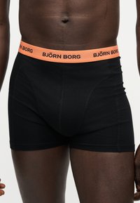 Ανδρικός κορμός που φοράει μαύρα μποξεράκια με πορτοκαλί μέση με ετικέτα "BJÖRN BORG" σε λευκό φόντο.