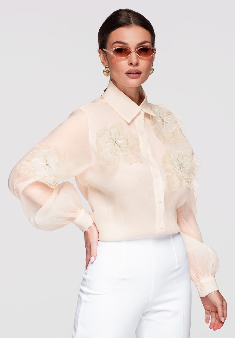 Femme portant un chemisier beige transparent avec des appliques florales, un pantalon blanc taille haute, des boucles d'oreilles dorées et des lunettes de soleil marron, posant la main sur la hanche.