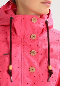 Giacca impermeabile rosa con pois neri, dotata di collo alto, bottoni in legno e lacci marroni con dettagli in legno.