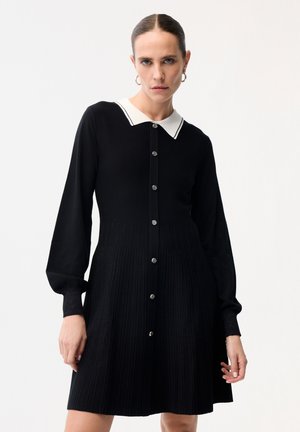 Frau mit zurückgebundenem Haar, die ein langärmliges schwarzes Kleid mit weißem Kragen und Knöpfen vorne trägt, steht vor einem schlichten hellen Hintergrund.