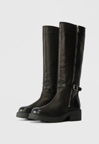 Schwarze knielange Lederstiefel mit seitlichem Reißverschluss, einer Schnallenakzent und einer robusten, strukturierten Sohle. Glatte Oberfläche und stabile Konstruktion.