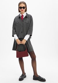 Grau kariertes Sakko mit kurzen Ärmeln und Knopfverschluss, weißes Hemd, rote Krawatte, grauer Rock, schwarze Slipper und eine rote Handtasche.