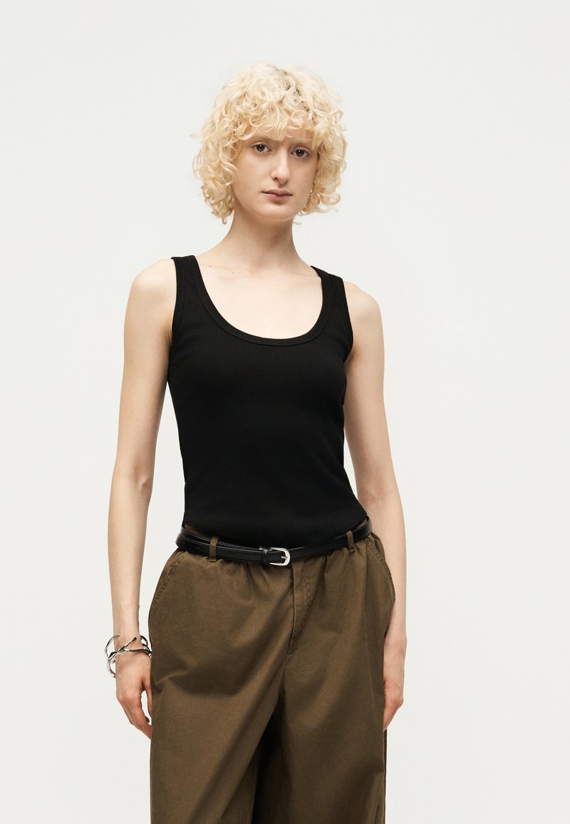 Άτομο με σγουρά ξανθά μαλλιά που φοράει μαύρο αμάνικο τοπ, καφέ παντελόνια με μαύρη ζώνη και ασημένιο βραχιόλι, στέκεται μπροστά σε απλό φόντο.