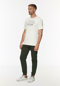 Witte t-shirt met korte mouwen en zwarte opdruk, gecombineerd met olijfgroene tapered broek en witte sneakers met elastische manchetten.