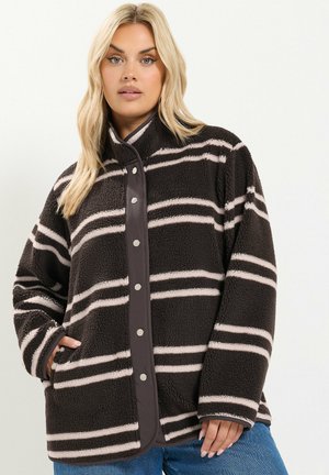 Femme portant une veste polaire rayée noire et beige avec des boutons-pression et des poches latérales, posant sur un fond clair uni.